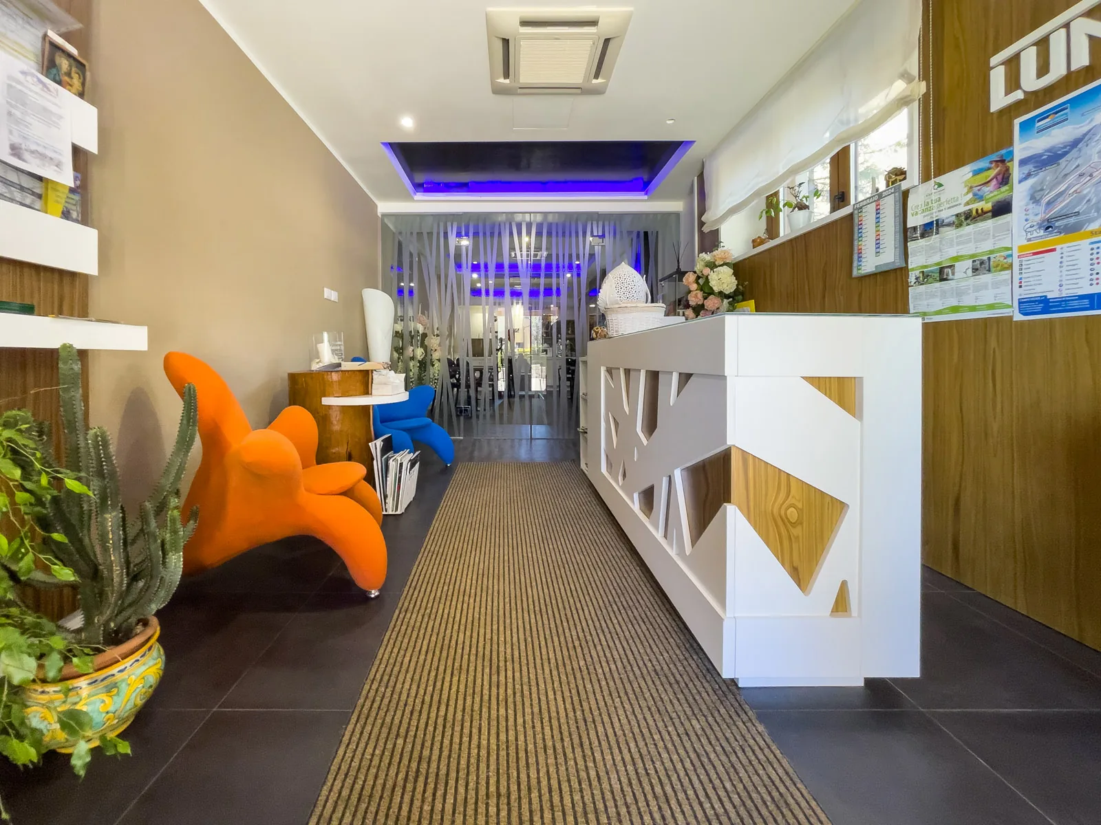 ingresso con bancone bianco traforato, poltrona arancione e piante, corridoio con tappeto verso la sala