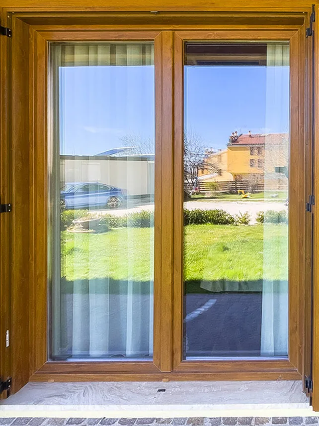 finestra con infissi in legno e tende bianche che riflette prato, strada e case